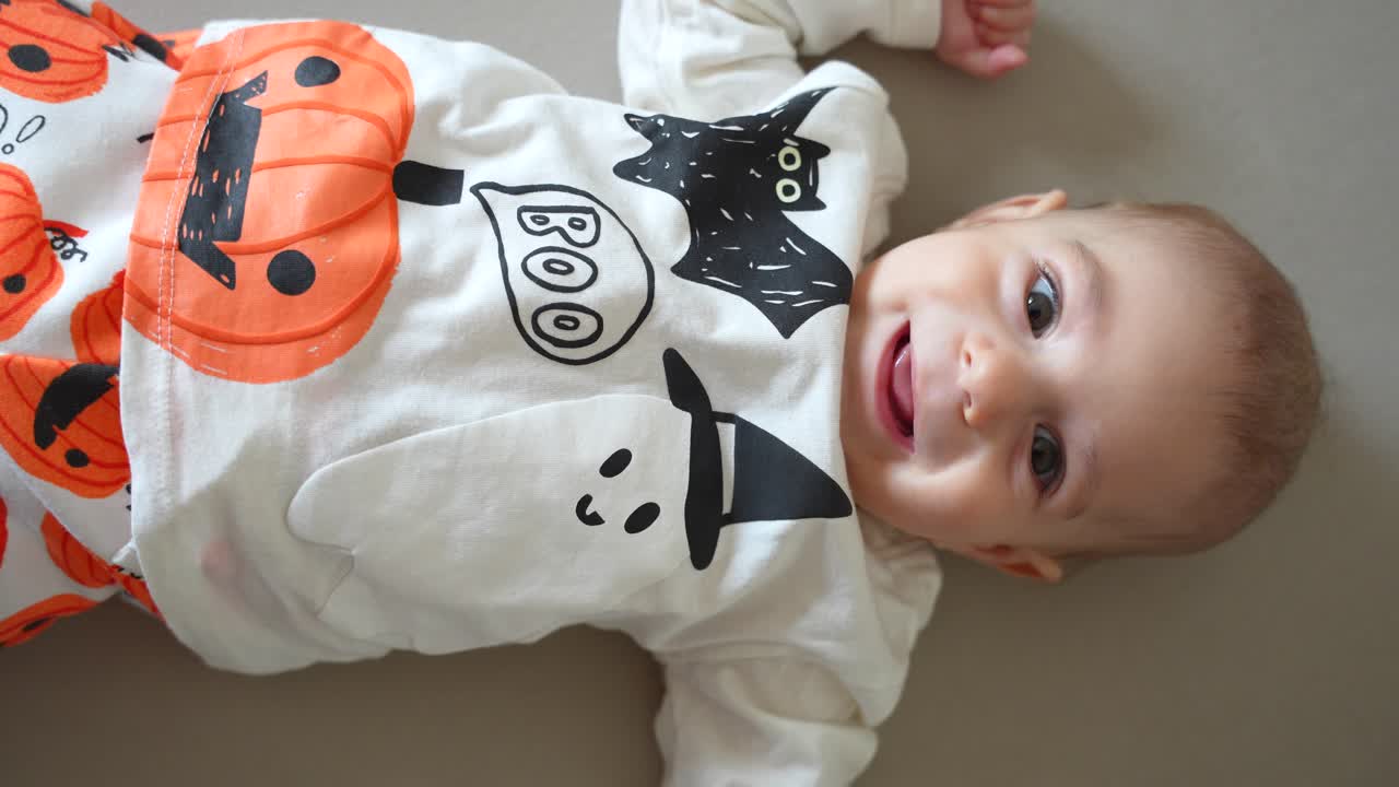 Happy 4 months old baby lying down smiling while looking at camera and wearing a Halloween jumper and pants with pumpkins, ghosts and bats