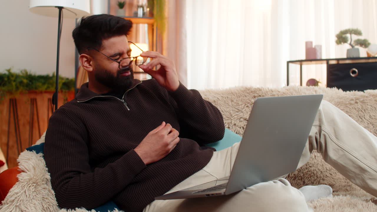 Indian man rubs tired eyes while on sofa with laptop at home eyes strained after long screen time