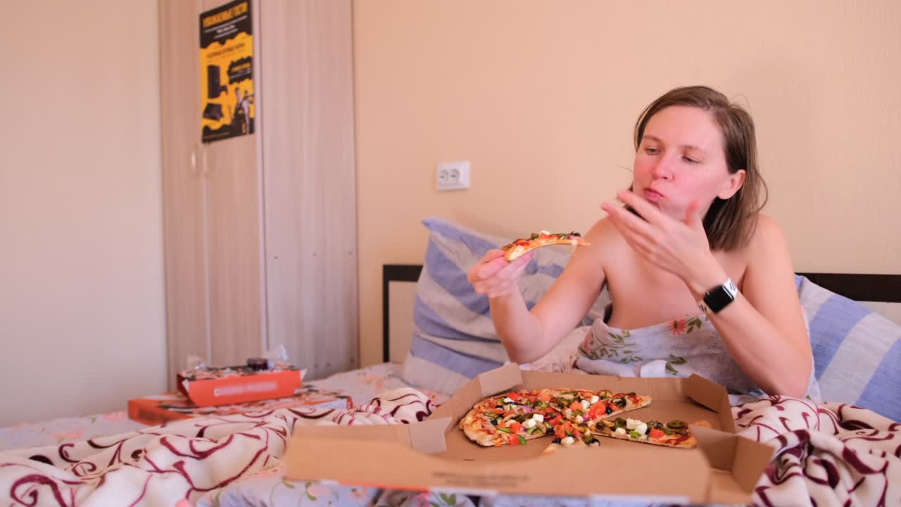 The girl lies in bed covered with blankets and eats delicious pizza in the bedroom with pleasure.