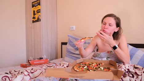 the girl lies in bed covered with blankets and eats delicious pizza in the bedroom with pleasure.