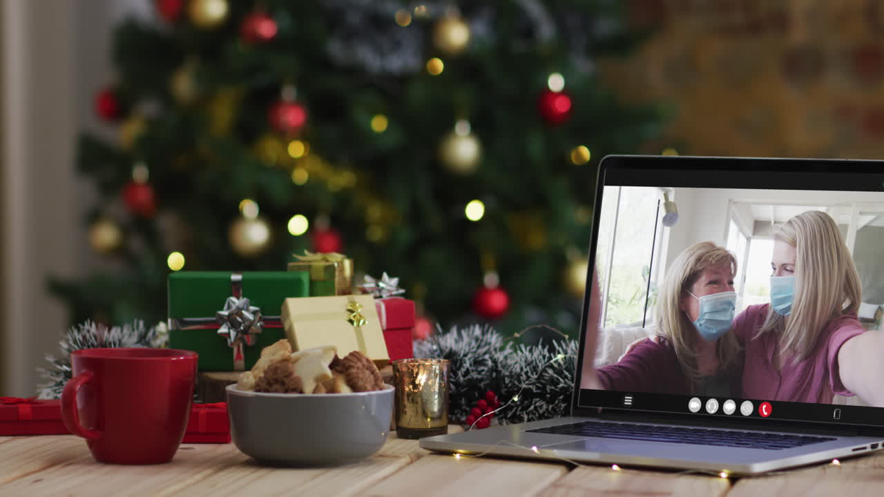madre e hija con máscaras faciales en una videollamada en una computadora portátil, con árbol de navidad