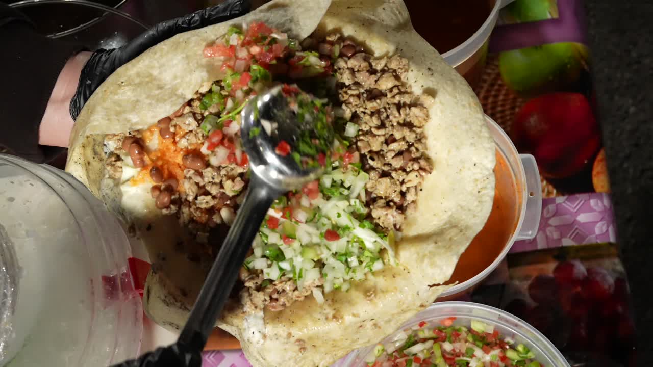 Nærbillede af en stor ske, der tilføjer frisk pico de gallo salsa til en fyldt mexicansk burrito. Fremhæver smag, tekstur og tilberedning ved en taco-bod i gadestil.