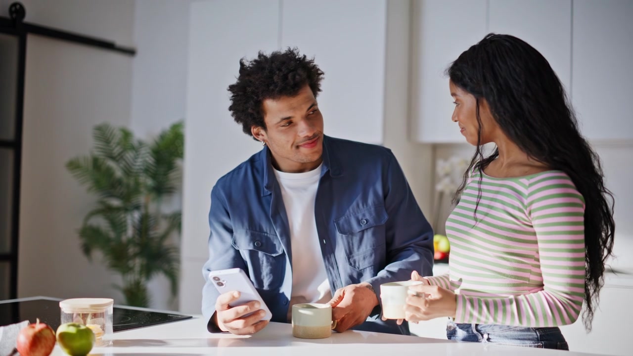 Smiling wife bringing coffee to husband holding smartphone in kitchen closeup