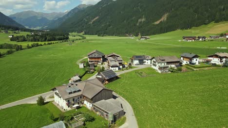 an alpine village is nestled among lush green fields and surrounded by forested mountains. traditional houses with solar panels and gardens are scattered across the landscape.