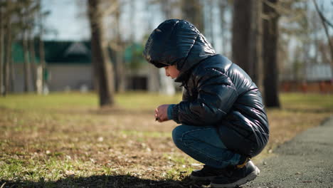 rotational view of young boy squats in a park, intently observing a stick in his hand, he is dressed in a black leather jacket, head warmer, and jeans, with a blurred background of trees