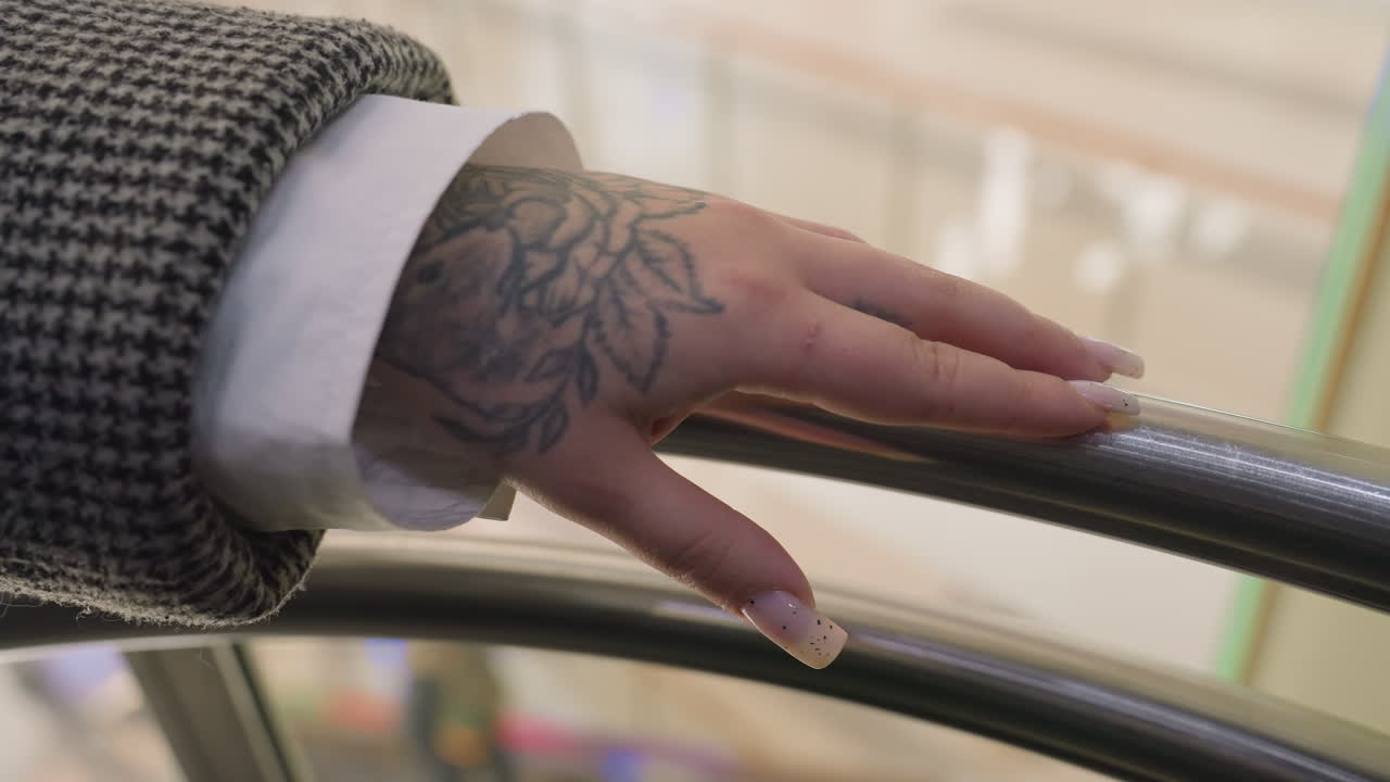 close up of tattooed hand gently tapping polished iron rail with soft background view seen through transparent panel inside descending elevator