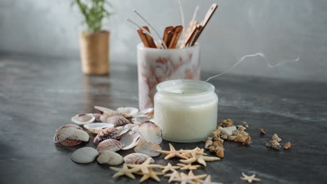 white candle releasing smoke after being extinguished, surrounded by seashells, starfish, and snail shells on marble table, with natural soft lighting