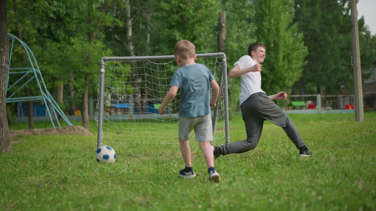 dos hermanos jugando al fútbol en un campo cubierto de hierba, el hermano menor patea la pelota hacia el poste de la portería y la persigue con entusiasmo, con ambos riendo