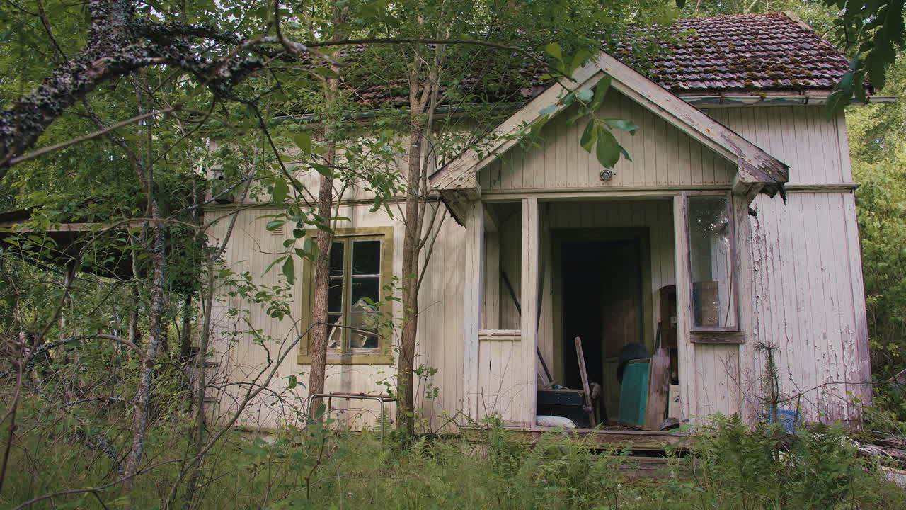 Abandoned house with peeling paint, broken windows, and dense overgrown vegetation.
