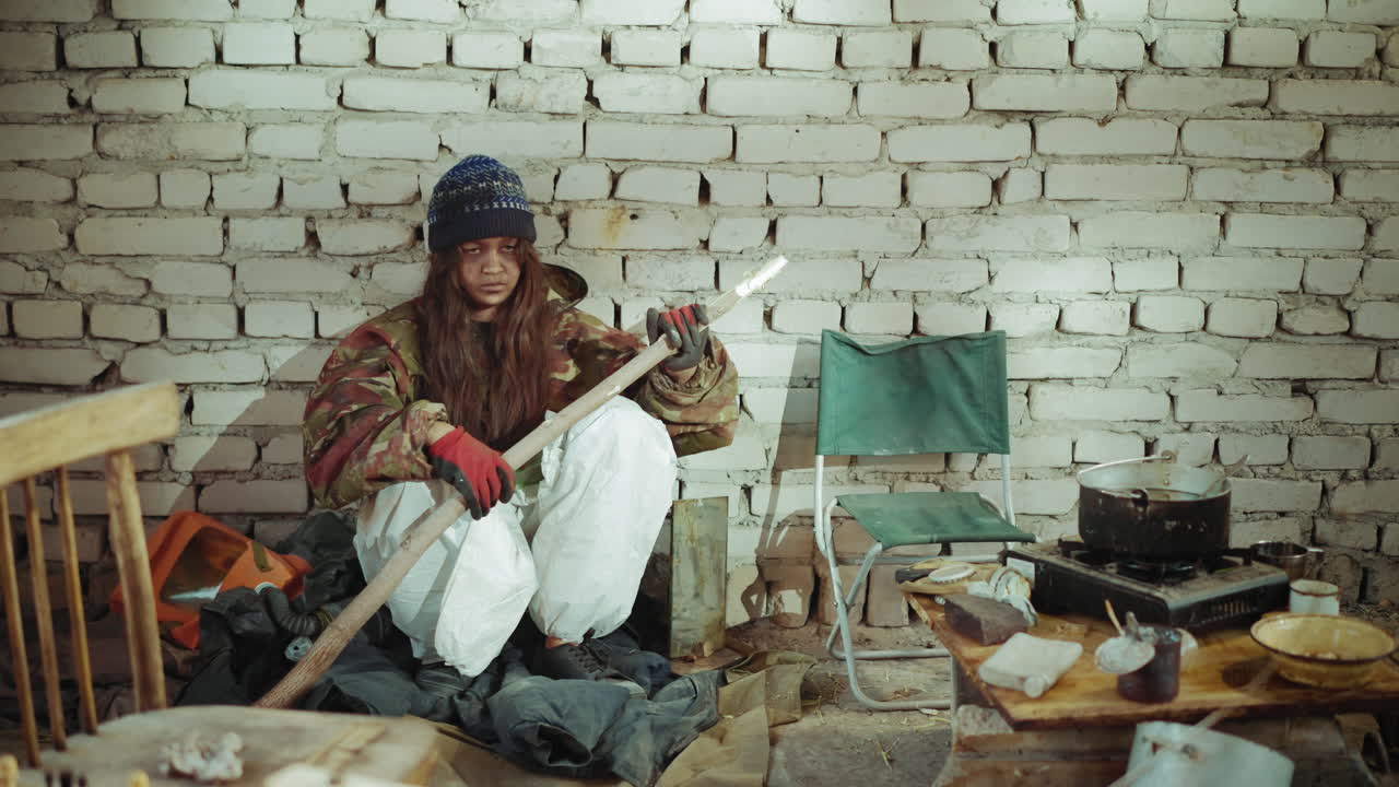 Orphan in military clothes sits against brick wall holding stick with tense expression, surrounded by makeshift cooking area and scattered belongings, symbolizing hardship