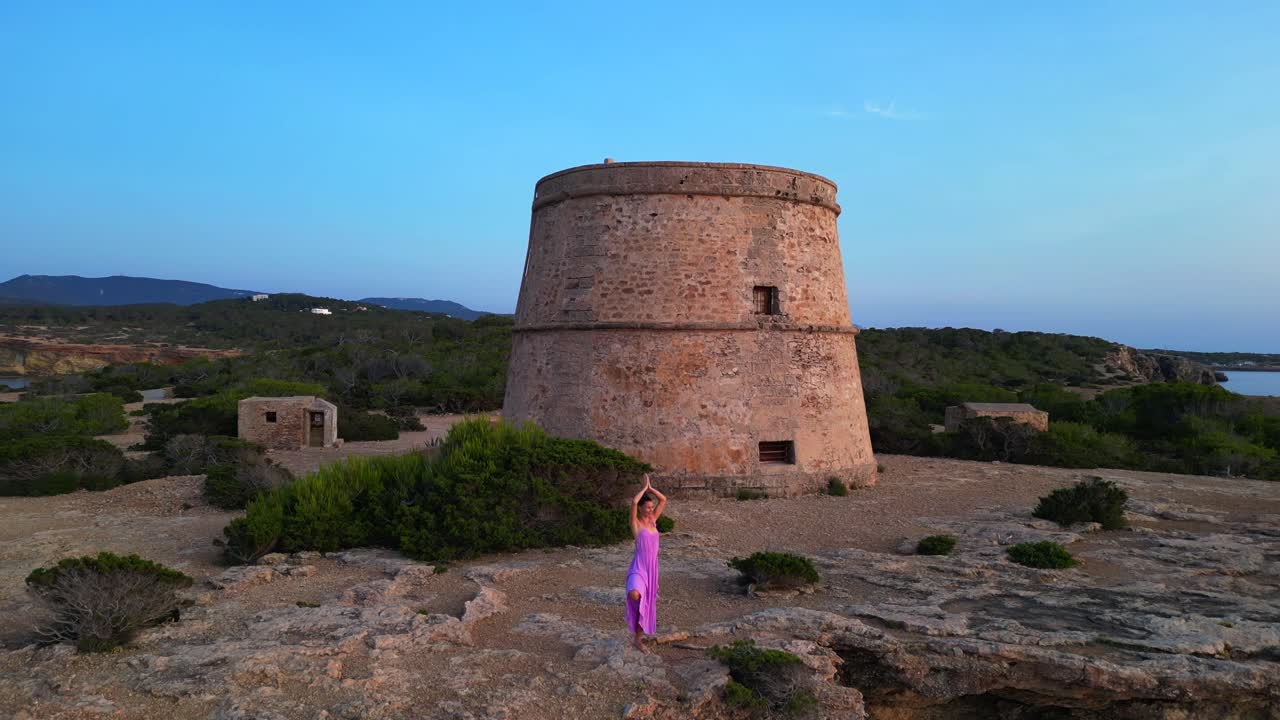 Woman in yoga pose enjoying the sunset near Torre d'en Rovira, a defense tower built in the 18th century in Cala Comte, Ibiza, Spain. Amazing aerial view flight speed ramp hyper motion time lapse