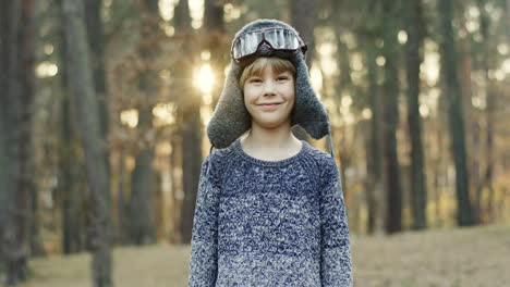 Little-Caucasian-boy-dreamer-of-being-a-pilot-having-fun-and-running-in-hat-and-special-glasses-smiling-at-the-camera-in-the-forest