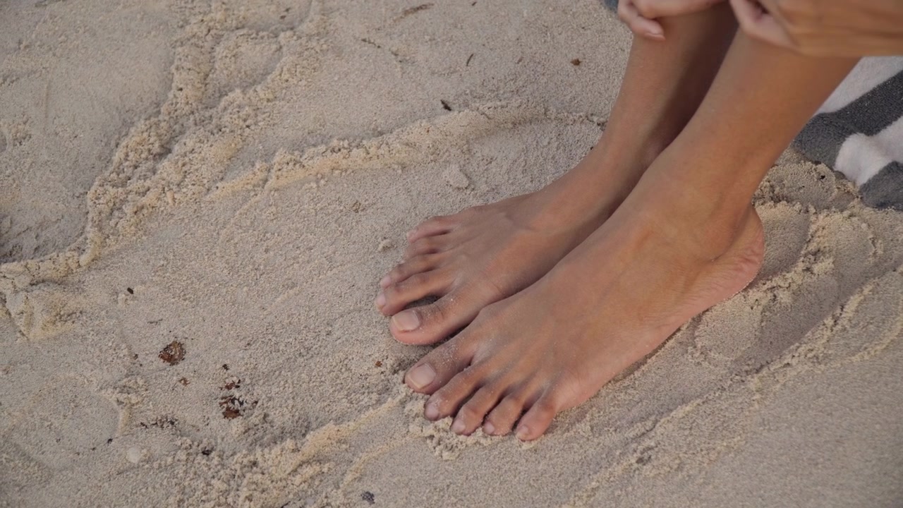 Mani che massaggiano piedi sabbiosi in spiaggia, delicato rituale di cura della pelle con braccialetto e asciugamano, granelli che scivolano tra le dita dei piedi, bagliore serale, movimento tattile ravvicinato, atmosfera rilassante di benessere e litorale tranquillo.
