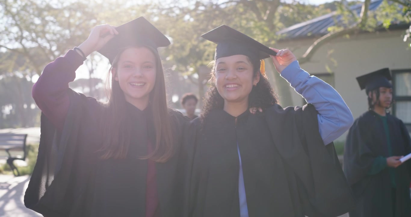Due studentesse diplomate adolescenti si abbracciano, si aggiustano i cappelli e posano in un cortile soleggiato festeggiando.