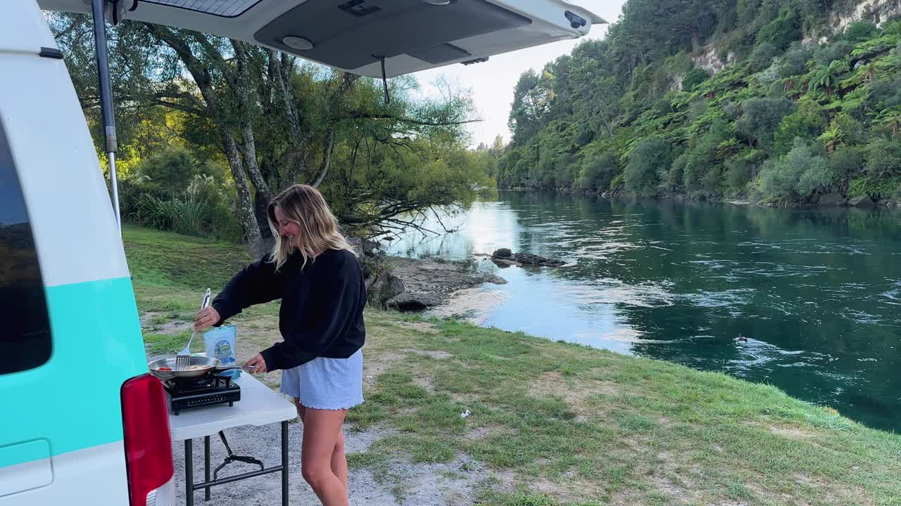 A woman cooks breakfast at the back of her camper van at Hipapatua Campsite, New Zealand. A peaceful morning surrounded by nature, outdoor living, and the freedom of van life travel