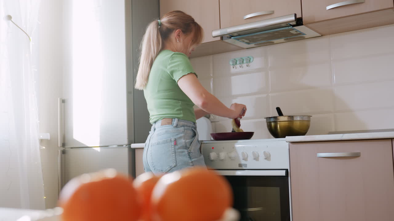 Perdeden güneş ışığı girerken yeşil tişörtlü bir kadın ocakta kızarmış yumurtayı çevirerek yemek yaparken aydınlık mutfakta tabaktaki taze portakalların yakın çekimi