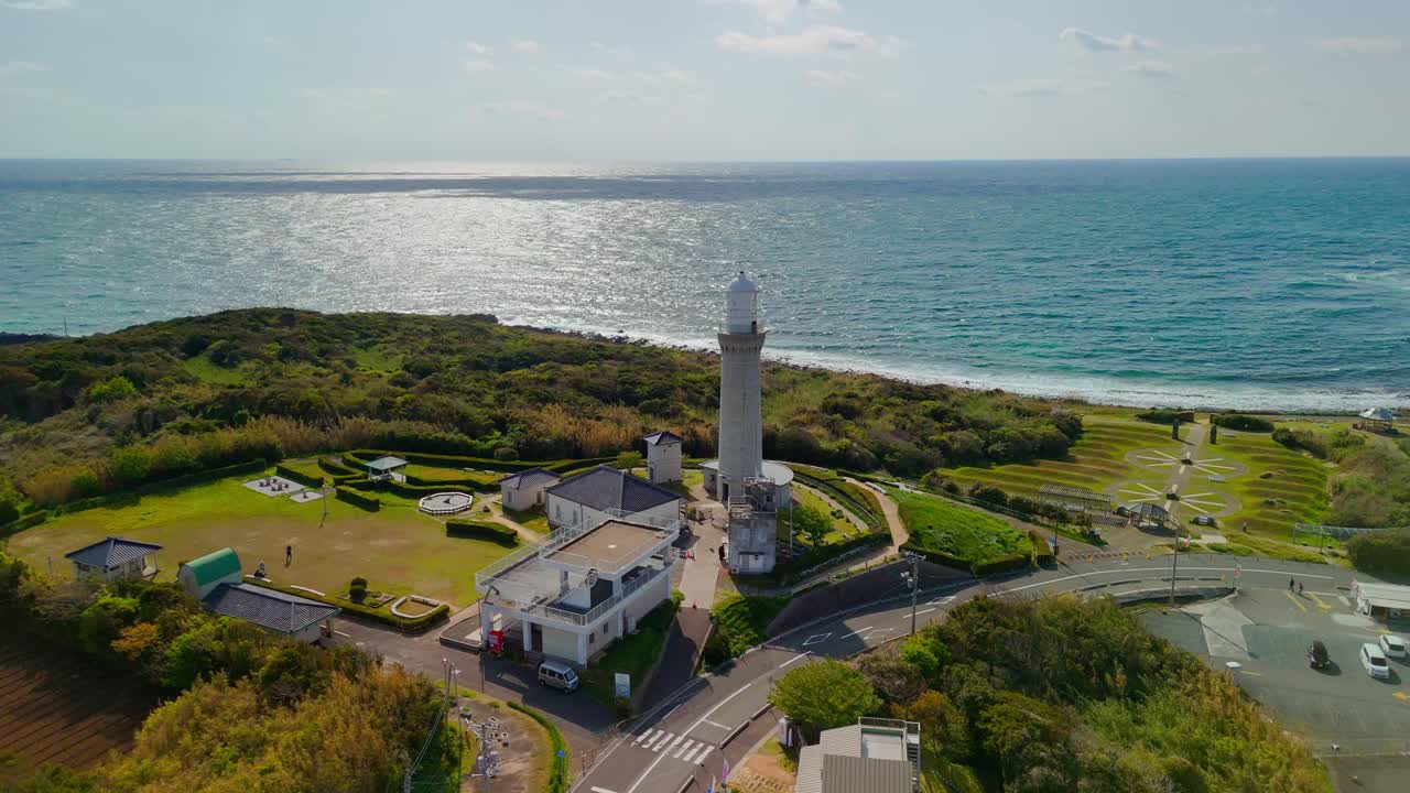 A beautiful aerial pan captures a lighthouse and visitor complex on a lush green peninsula in Japan, with the sun reflecting brightly on the vast ocean