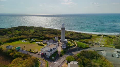 a beautiful aerial pan captures a lighthouse and visitor complex on a lush green peninsula in japan, with the sun reflecting brightly on the vast ocean