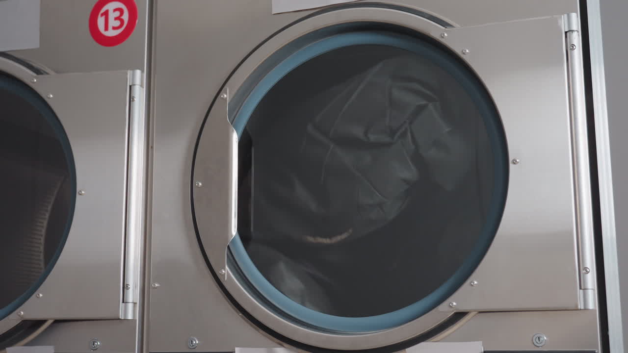 Front load industrial washing machine in operation with fabric rolling inside drum behind round glass door, stainless housing in laundromat, cycle running, close up spin and motion, cleaning process