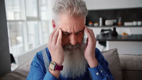 primer plano de un anciano con cabello gris y barba llena en una camisa azul amasa y masajea sus sienes durante un dolor de cabeza en un apartamento moderno