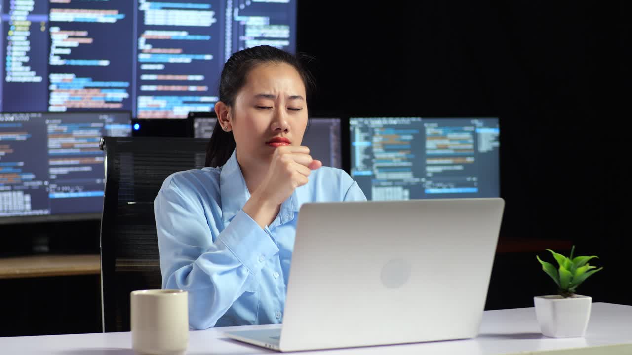 Asian Female Programmer Yawning While Writing Code By A Laptop Using Multiple Monitors Showing Database On Terminal Window Desktops In The Office