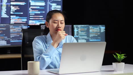 asian female programmer yawning while writing code by a laptop using multiple monitors showing database on terminal window desktops in the office