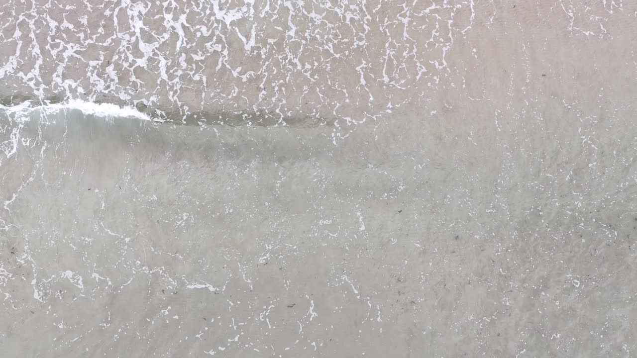Top-down drone shot of calm waves creating white foam on sandy beach, soft natural lighting