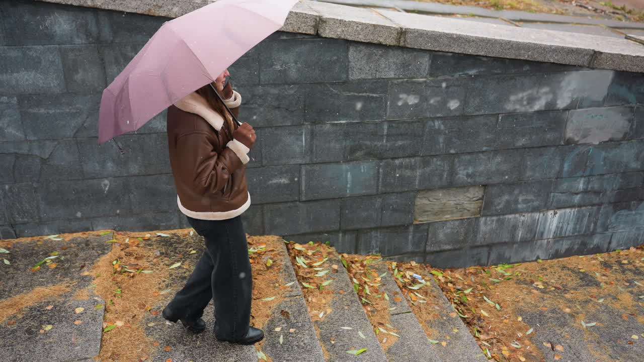Up shot of young girl smiling on phone call holding umbrella, wearing knit cap, brown shearling jacket, black trousers, walking down stone steps during light snowfall with colorful autumn leaves