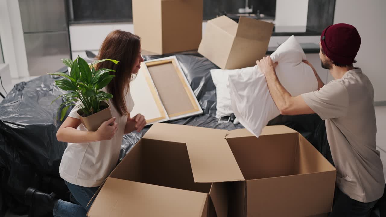 Happy brunette girl in a white T-shirt holds a house plant in her hands and takes items out of a box together with her boyfriend in a beige T-shirt during her move to a new apartment