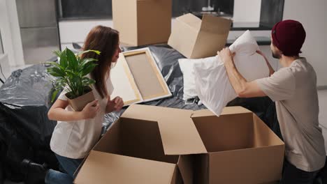 Happy-brunette-girl-in-a-white-T-shirt-holds-a-house-plant-in-her-hands-and-takes-items-out-of-a-box-together-with-her-boyfriend-in-a-beige-T-shirt-during-her-move-to-a-new-apartment