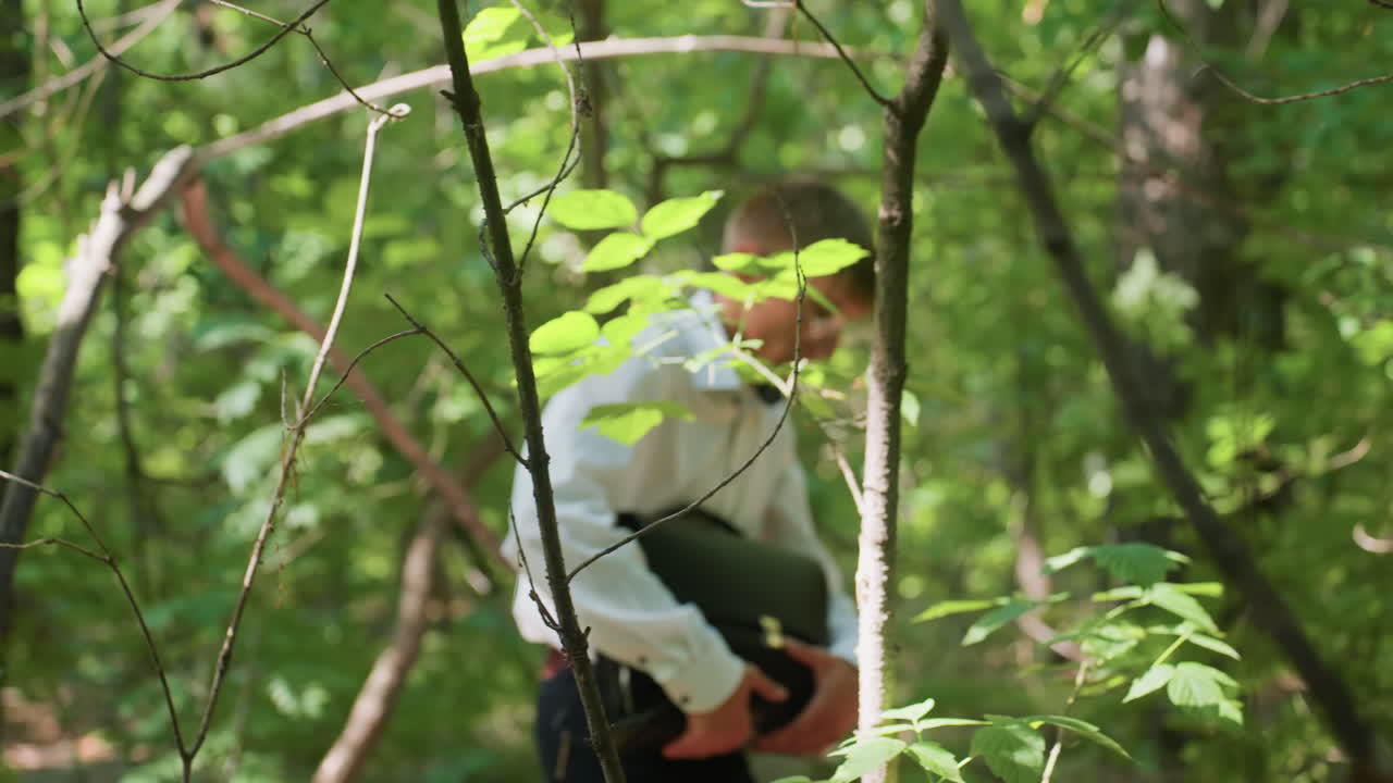 Side view of biologist in white shirt carrying backpack while bending carefully under branches in dense green forest, moving with focus and caution through natural woodland during daylight