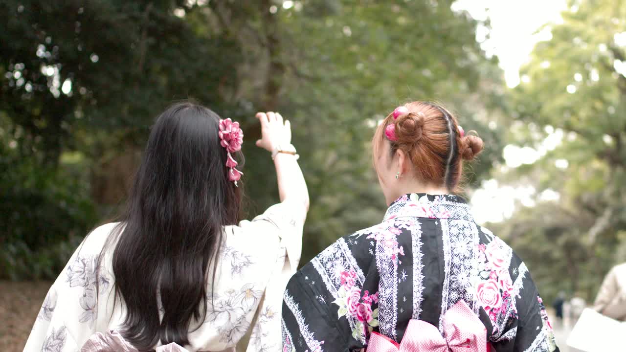 Cảnh quay từ phía sau hai người phụ nữ mặc yukata họa tiết hoa đang đi bộ qua một công viên xanh tươi