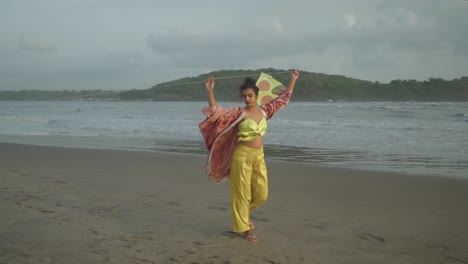 a woman with yellow clothes holding a kite and walking alongside the beach