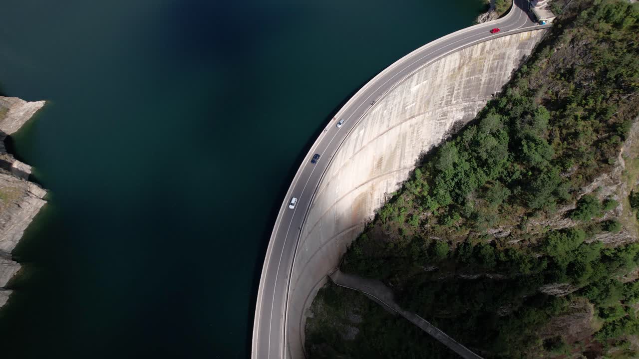 Aerial shot of cars driving on the Vidraru Dam in Romania, circling left. Blue lake, dramatic curves, and green mountain backdrop in 4K.