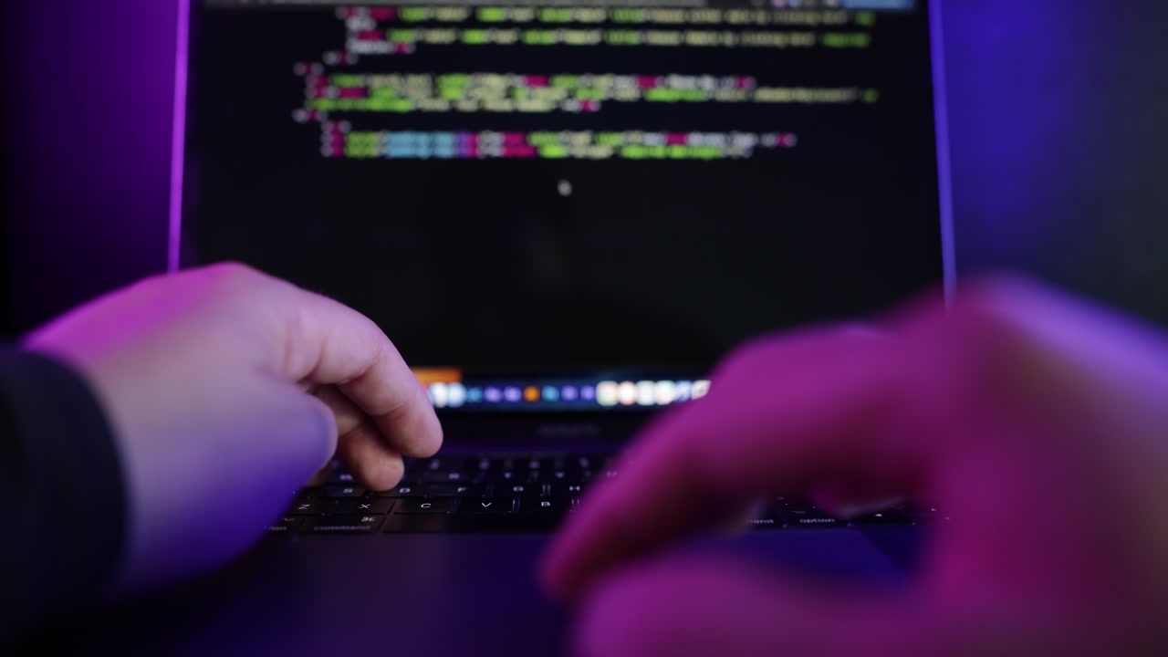 Dark cinematic close-up footage of software developer coding on a laptop in a modern neon-lit environment with blue and purple ambient lighting.