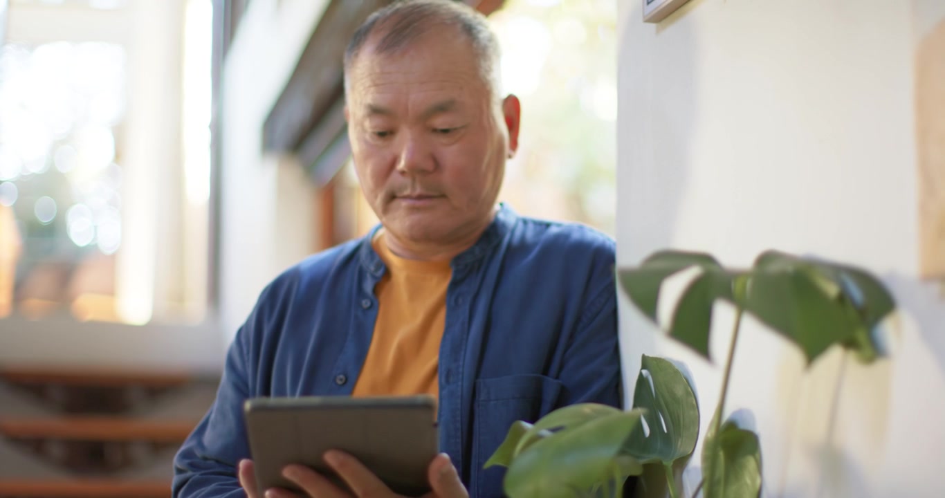 窓際の植物のそばで、タブレットを持ち、画面をタップしたりスワイプしたりしながら自宅で読書するアジア系の高齢男性