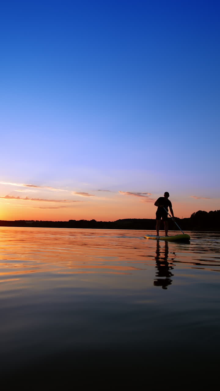 Sportsman practices paddle boarding on the river at sunset. Man stands on the sup board moving with an oar. Vertical video.