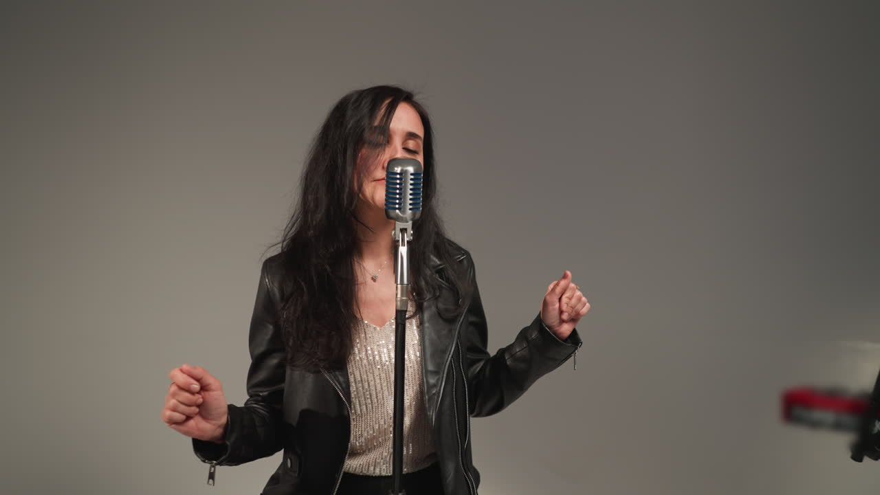 A vocalist in a black leather jacket sings with expressive hands and eyes open, standing in front of a vintage microphone against a grey background