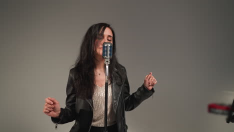 a vocalist in a black leather jacket sings with expressive hands and eyes open, standing in front of a vintage microphone against a grey background