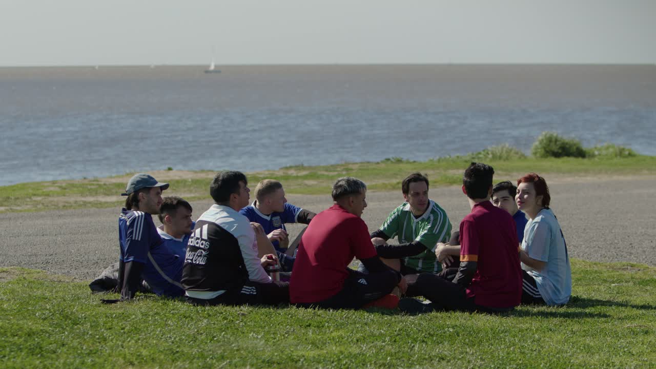 A group of football soccer players sit on grass near the ocean, talking during a break in a game