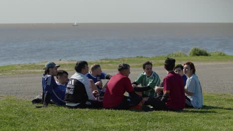 a group of football soccer players sit on grass near the ocean, talking during a break in a game