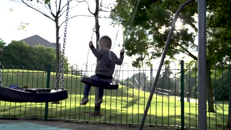 three year old boy being pushed on swing in late afternoon summer sun setting behind him, in playground, uk