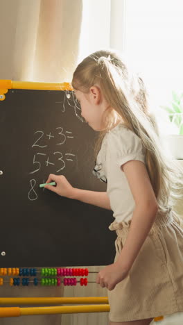 Little schoolgirl writes problems on blackboard and looks around at brother playing school at home slow motion. Boy practices math sitting at desk closeup