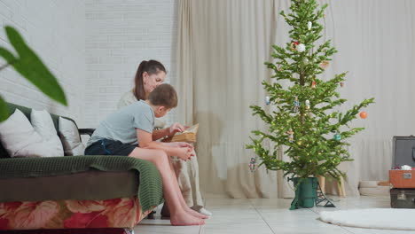 side view of boy in grey shirt and black shorts sitting on couch with mom in brown gown reading old book near decorated christmas tree with cotton ornaments cozy indoor family bonding moment