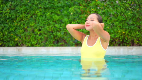 Beautiful-Thai-young-woman-in-pool-with-yellow-swimsuit-strokes-wet-hair