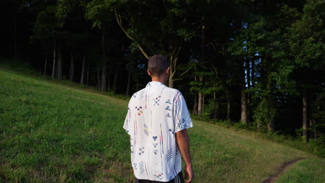 hombre joven con camisa y pantalón corto camina descalzo por un pequeño sendero en cámara lenta hacia un bosque verde en un día soleado de verano