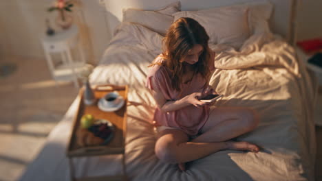 smiling woman messaging online in cozy bed. carefree girl typing mobile phone