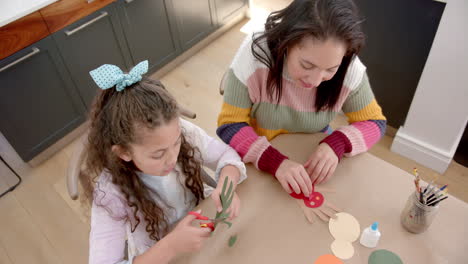 Happy-biracial-mother-and-daughter-playing-with-paper,-talking-and-smiling-in-sunny-living-room