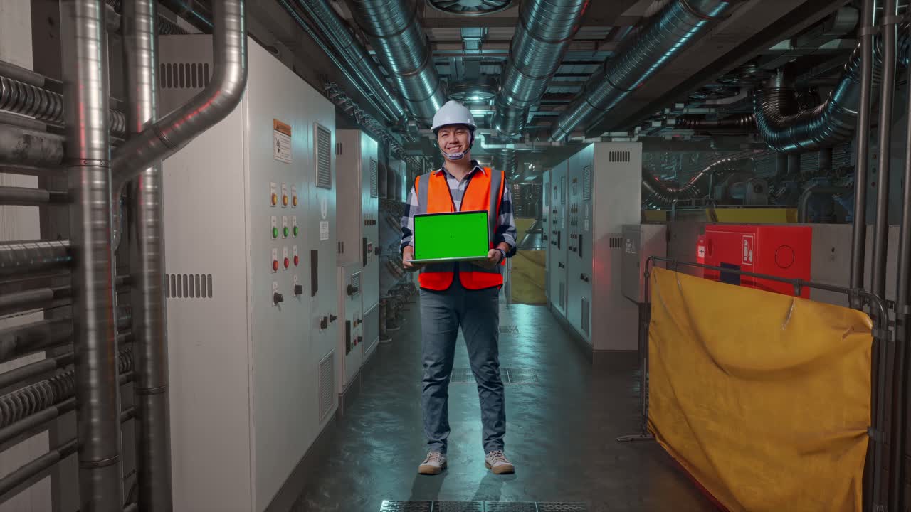 Full Body Of Asian Male Engineer With Safety Helmet Smiling And Showing Green Screen Laptop To The Camera While Standing In Engine Control Room, Work Of Electrical Generators