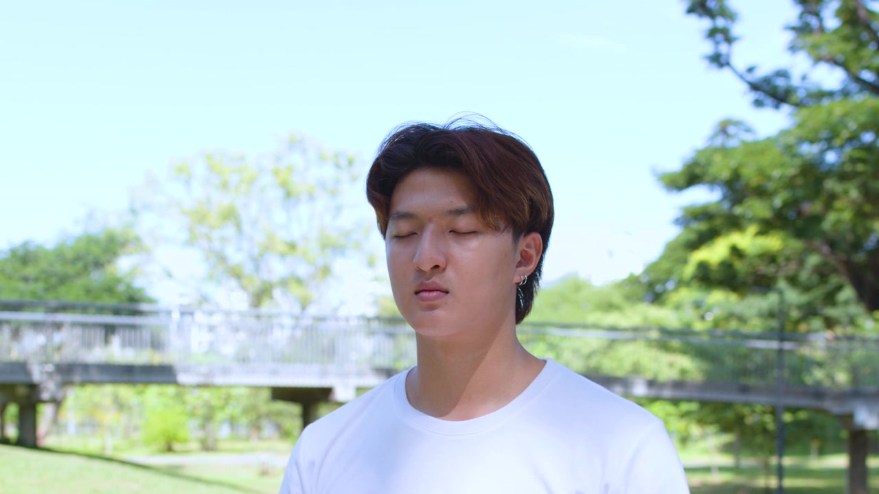 若いアジア人男性が、豊かな緑に囲まれた日当たりの良い公園で、屋外でマインドフルネス瞑想を実践している。